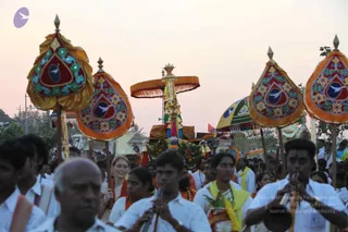 Evening procession at dusk with a crowd of devotees in white shirts and dhotis, several with saffron and yellow shawls. Four tall ornate parasol-like standards with orange and red fringed canopies, decorated with floral and bird motifs, rise above the crowd. Central palanquin or ratha canopy with a saffron fabric top, tassels and garlands, carried through the street. A young man in the foreground plays a long wind instrument, and another in white plays cymbals. Several women in white and yellow saris walk among the procession. Warm pink sky and trees in the background.