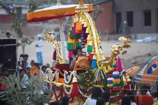 A photograph of a golden processional chariot (ratha) decorated with garlands of white and purple flowers, colourful tassels in green, pink, blue and yellow, and hanging bells, with a golden umbrella canopy and carved golden horse-head ornaments at the front. A person seated on the chariot wears an orange-gold shawl and raises one hand in greeting, while a crowd of onlookers, mostly men in white and bright clothing, lines the procession route around the vehicle. A flowered garland and a red-and-orange canopy frame the scene, with a loudspeaker on the left and low buildings in the background.