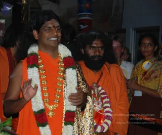 Photograph of a smiling man in orange robes wearing a long white and red flower garland with a heavy gold chain of ornaments and a leopard-print shawl, raising one hand in greeting. Beside him, a bearded man with long dark hair in orange robes holds a small bronze deity murti adorned with a pink and white flower garland. Behind them a woman in a yellow patterned sari with a yellow badge stands, and a person in white is partly visible. A red and white banner-like pillar and a red-and-yellow wall painting appear in the background. Visible physical people: four. The smiling man has a red tilak on his forehead.