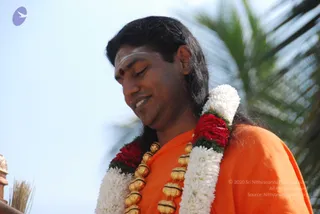 Close-up photo of a man with long swept-back black hair wearing an orange saffron robe and a layered garland of white chrysanthemums, red roses and gold-coloured beads around his neck. He has a white tilak mark and a red bindi on his forehead, eyes lowered and a faint smile. Blurred palm fronds and a pale blue sky fill the background, with a partial stone pillar visible at the lower left.