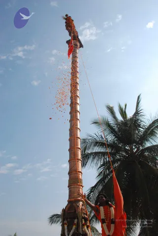 A tall copper flagstaff rises against a bright blue sky, topped with a carved finial and a red-orange flag, while orange flower petals scatter down its length. At the base, a man in orange robes and a white and orange garland raises one arm beside the pole, with a palm tree behind him and marigold garlands wrapped around the staff's base. A thin orange rope runs up the pole, consistent with a flag-hoisting ceremony.