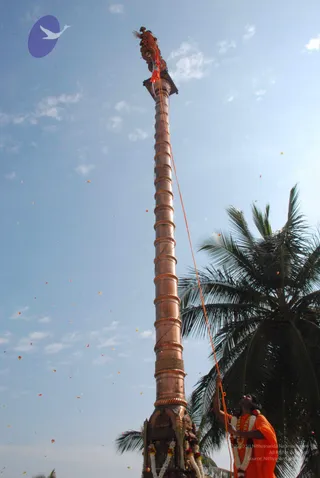 A tall copper flagstaff rises steeply against a pale blue sky, its top crowned with a decorated finial and a red-orange tassel. A man in orange robes and white-and-red flower garlands stands at the base, pulling an orange rope that runs up the pole, as he hoists the flag. Small orange petals or flower fragments drift through the air around the pole. Tall palm fronds appear at the right edge, and the base of the pole is decorated with marigold-style garlands.