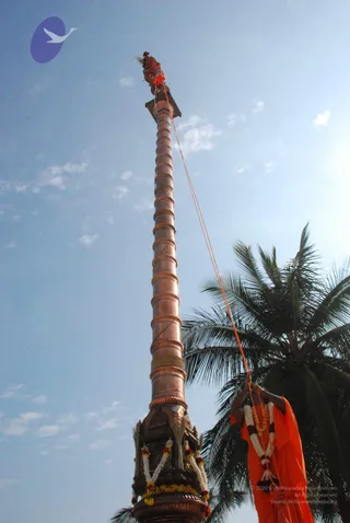 Low-angle photo of a tall copper flagstaff with spiral ridges rising against a bright blue sky, topped by a small ornate finial with an orange cloth. Its base is decorated with white and orange marigold garlands. A figure in an orange robe stands at right beside a coconut palm, holding an orange rope that runs up to the flagstaff, with a garland and beaded necklace visible at the chest. Wispy white clouds fill the sky.