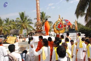 Outdoor flag-hoisting scene beside a tall copper flagstaff with a decorated base, set on a white platform among palm trees. A person in orange robes stands atop the platform beside the pole, which is garlanded with flowers. Nearby, a richly embroidered ceremonial canopy in gold, red and teal with a dove motif is held on poles. Around them, many devotees in white kurtas and dhotis wear yellow shawls and stand in a crowd, several with hands raised or pointing. Thatched huts and a palm grove form the background under a pale sky.