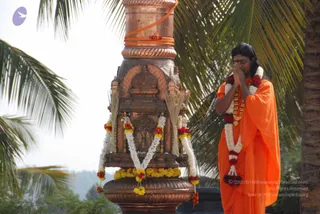 A man in orange saffron robes stands outdoors at right with his palms pressed together in namaskar, eyes lowered, wearing a red bindi, a white and red flower garland and a gold-toned necklace. Beside him is an ornate copper-coloured sculpted pillar or kalasha-style stand, wrapped with an orange cloth, decorated with marigold and white flower garlands, yellow marigold clusters at its base, and dry grass or palm fronds tucked at its sides. Palm trees and a hazy sky fill the background. Swamiji (probable): long swept-back dark hair, saffron robes and a rudraksha-style necklace indicate the Swamiji candidate, who is shown here in an in_person posture of devotion.