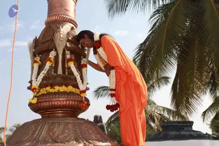 A man in a saffron-orange robe, wearing white and red-yellow flower garlands and a gold chain, bows forward with his forehead pressed against an ornate copper flagpole decorated with marigold and white flower garlands, and with dry palm-frond fans tied to its top. The flagpole base is carved with figures and sits on a stone platform. A coconut palm frames the right side under a pale sky, with a dark stone sculpture visible near the base. The pose suggests a devotional gesture at a flag-hoisting. No other people are visible.