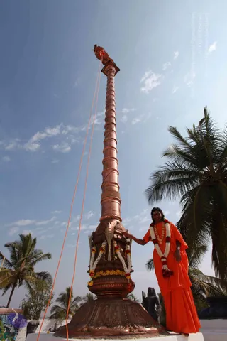 A tall, slender copper flagpole stands on an ornate copper-clad base decorated with floral garlands of marigolds and white flowers, with orange ropes running up from it into the sky. A person in a flowing saffron-orange robe with a multi-strand garland stands beside the pole, one hand resting on the base near the flower garlands. Coconut palms frame the open blue sky, and a dark stone sculpture is visible at the lower right. A partly visible sign lettering appears at the lower left. The pole is the central element of a flag-hoisting ceremony.