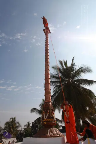 Photograph of a tall copper flagstaff with a spiral-banded shaft rising against a pale blue sky, topped with a small red-and-gold figure and a red flag. A saffron-robed person with a white flower garland stands at the base pulling a red rope that runs up the pole, while a second person in orange clothing and a third in white kneel beside the base. The base is wrapped in marigold and white flower garlands and sits on a white platform, with coconut palms behind and a dark stone sculpture at lower left.