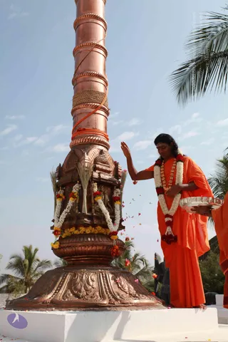 A man in a saffron-orange robe with long dark hair stands on a white platform beside a tall copper flagstaff. The base of the pole is a decorated copper shrine with carved details, draped with white and red-and-yellow flower garlands and bundles of dried grass. He wears a white and red flower garland and a wooden bead necklace, and with one hand he sprinkles petals from a silver tray held by another robed attendant at the right edge. Palm trees and a bright blue sky surround the scene.