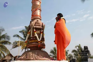 A figure in flowing saffron-orange robes stands with palms pressed together in prayer beside a tall copper flagpole mounted on an ornate copper base. The base is decorated with carved panels and a small shrine niche holding a seated deity murti, garlanded with marigold and white flower strands, with bundles of straw and orange cloth wrapped around the pole. Scattered petals lie on the platform. Palm trees and a clear pale sky surround the scene, and two dark stone sculptures stand at the right edge. The figure has long dark hair and wears a white flower garland, and the scene suggests a flag-hoisting ritual at an outdoor ashram setting.