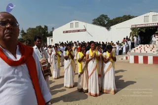 Outdoor photo at a temple compound. In the foreground, a bald man with glasses and a red shawl over white clothes stands facing the viewer. Behind him, a line of women in white saris with gold borders and yellow shawls stand with palms pressed together in namaste, eyes lowered. A drummer with a barrel drum stands among them. Behind, a crowd in white clothes gathers before a whitewashed temple with red-and-white steps and a signboard reading Sri Anandeshwara Mandir; another sign on the right reads Sri Anandeshwara. No a person candidate is clearly visible.