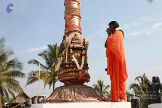 A person in a flowing saffron-orange robe stands with palms pressed together in prayer beside a tall copper flagstaff-like pillar. The pillar is wrapped with a braided rope and an orange cord, and its base is decorated with marigold and white flower garlands, red blossoms, and scattered petals on a white platform. A small golden deity figure sits in a niche on the pillar. Palm trees and a blue sky surround the scene, with dark stone sculptures visible at the right edge. The person has a white garland around the neck and is shown in profile with eyes closed, appearing to be a sannyasi-style devotee during a flag-hoisting ceremony.