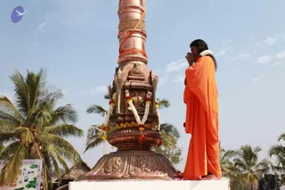 A photograph of a single person standing with hands pressed together in namaste before a tall copper flagstaff on a white raised platform. The person wears an orange saffron robe draped over the shoulders and has long dark hair, with a white flower garland around the neck. The flagstaff is wrapped with an orange cord and topped with a copper shrine-like base holding a small seated deity murti, decorated with marigold and white flower garlands, yellow flowers and red blossoms scattered on the platform. Coconut palms and a blue sky with clouds form the background, with a green map sign at the lower left. Swamiji (probable) is shown here by appearance cues: long hair, orange robe and garland, consistent with the page's reference to Swamiji at the flag-hoisting event; the identification is based on those cues alone.