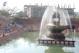 A photograph of a large black stone linga on a raised platform in the middle of a water-filled pool, with fountain jets of water spraying up around it. The linga is decorated with a yellow and red floral garland band and a flower garland around its base. Tiered red-brown stone steps on the left hold a seated group of people in mostly white and saffron clothing, facing the linga, while a partially built multi-storey concrete building with exposed rebar stands behind the site and bare trees sit at the left edge. No a person candidate is visible in the scene.