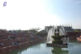 A large outdoor photo of a water-fed reflecting pool with a black Shiva linga on a square base, garlanded with yellow and gold festoons, with water jets spraying up around it. Tiered red steps on the left and right hold many seated and standing people, mostly in white clothes, some with red and orange garments. A partly built multi-storey concrete structure with rising rods stands in the background under a pale sky. Some people walk along the steps and a few sit at the water's edge.
