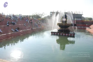 A large black linga stands on a square platform in the middle of a rectangular reflecting pool, with water jets spraying up around it. The linga is decorated with a band of colourful flower garlands near its base. Red-tiered stone steps on the left side of the pool hold a crowd of people, many dressed in white and some in orange or coloured saris, seated and standing in clusters. Unfinished concrete buildings with rebar rise in the background under a hazy white sky.