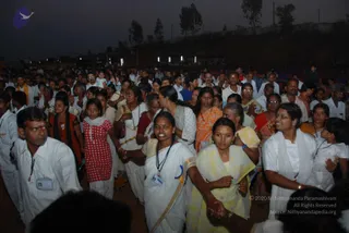 Dusk outdoor gathering of a large crowd of devotees, mostly in white clothing, standing closely together and holding hands in lines. Many wear white shirts, white dhotis, white saris and white salwar kameez, with some in orange, yellow, red and pink garments. Several women wear necklaces and beaded malas, and a few men wear lanyards with identity badges. A woman in a yellow sari and a woman in a white sari hold hands at the centre-front, and a man in a white shirt with a badge stands at the left. Floodlights and a hillside with trees form the background.