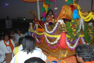 Outdoor evening procession with a long-haired figure seated on an ornate golden chariot (ratha) draped in marigold, white, purple and yellow garlands, with green foliage, red and blue tassels and a golden kalasha-topped canopy. The seated person wears an orange-saffron shawl and white garment, smiles and raises a hand in greeting; the figure has long swept-back hair and a white flower garland, consistent with the Swamiji (probable) cues of long hair and saffron wrap. Around the chariot, devotees in white and yellow dhotis and saffron scarves walk and crowd close, one man in a white shirt and orange shawl wearing a mala at left. Dusk sky and a stage structure in the background.