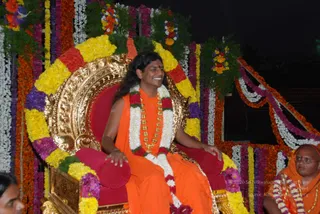 Photo of a man seated on an ornate gold-and-red throne decorated with yellow, orange, purple and white flower garlands, at night. He wears an orange saffron robe, a long white-and-red flower garland and a gold bead necklace, with long dark hair swept back, smiling with his hands resting on the red cushioned armrests. Behind him are more flower garlands and a dark night backdrop. To the right, a bespectacled man in orange robes and a flower garland sits partly visible. Swamiji (probable) is seated on the throne, with long swept-back hair, saffron robe, and ceremonial gold and flower adornments. A partially visible person stands at the far left edge.