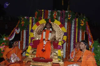 A central figure sits on an ornate gold throne with red cushions, raising one hand in a greeting or blessing while smiling. He wears a saffron-orange robe, a white and red flower garland, a beaded rudraksha-style mala and a gold necklace, with a leopard-print cloth draped over the throne. The throne is framed by a dense arch of marigolds and roses in yellow, orange, pink, purple and white, with greenery and garlands. Two attendants in orange robes and gold necklaces sit on the lower platform, one to the left gesturing toward the throne and two to the right, one bald and one with long hair. The scene is set at night with a dark background.
