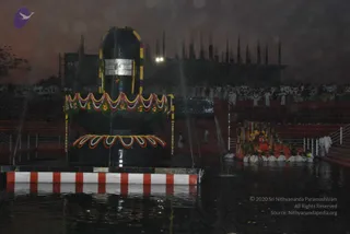 A large black Shiva linga stands in a shallow water pool at night, decorated with a silver-framed front face and a garland of multicoloured flowers wrapped around its base, with a floral ring at the bottom. Water sprays from fountains around it. To the right, a decorated boat or palanquin carries figures in orange and red robes, and several people in white clothing stand in the water beside it. Behind, a large crowd in white stands in tiered stands, with a building lit against the dark sky. The scene suggests a night abhishekam at a linga, with many white-clad participants.