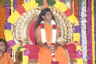 A photograph of a long-haired man in saffron-orange robes seated on an ornate gilded throne with red cushions, framed by marigold and yellow garlands and flower strands. He wears a white and red flower garland, a gold bead necklace, and a red bindi on his forehead, smiling and gazing upward with hands clasped. Beside the throne at left, a second man in orange robes with a gold necklace looks to the side. Swamiji (probable): the long swept-back hair, saffron clothing, ornate gold necklace and seated throne position are distinctive visible cues for the central seated figure.