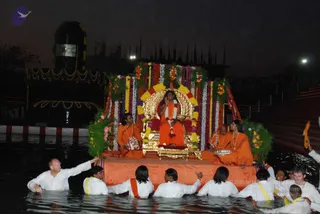 Night photo of a floating golden platform on dark water, decorated with garlands of marigolds and white flowers. A central figure in orange robes and a rudraksha mala sits on an ornate golden throne-like seat with a red cushion, raising one hand, while two attendants in orange sit on the platform beside him. White-clad devotees wearing yellow sashes stand waist-deep in the water pushing or steadying the platform. A dark stone shivalinga-shaped structure with silver bands and illuminated lamps stands on the shore behind, with a lit staircase at right. Swamiji (probable): the seated central figure wears orange robes and a rudraksha mala, with long dark hair swept back, consistent with the page's reference to Swamiji at this event.