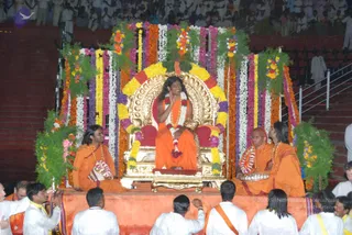 A photograph of an outdoor evening event on a raised orange stage in front of a large seated crowd. A central figure sits on an ornate gold throne with a red cushion, wearing an orange robe, a white flower garland and a red-orange garland, and a long-haired appearance; the throne is framed by tall arrangements of marigolds, yellow, white and purple flowers and green foliage. Two orange-robed attendants sit on the stage beside the throne, one with long hair and a flower garland holding a cloth-wrapped object, the other holding a silver tray with garlands. Men in white clothes and orange shawls kneel and sit at the front of the stage, and audience members fill the stands behind. Whether the central figure is the subject of the page cannot be confirmed from appearance alone, so the identity is marked unsure.