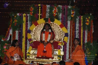 A seated man in saffron-orange robes sits on an ornate gold throne with a red cushion, framed by a dense arch of marigold and mixed flower garlands in yellow, orange, purple and white. A white and red floral garland hangs around his neck, with a long gold-and-rudraksha style necklace and a leopard-print cloth draped over the armrest. Two attendants in orange robes sit on either side at the foot of the stage, one holding a small floral offering tray, and strings of lights glow in the background. Several rudraksha-style beaded necklaces and garlands are visible. The setting appears to be a ceremonial stage decorated for a Shivaratri-type celebration.