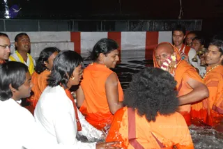 A photograph of a group of devotees standing waist-deep in dark water beside a red-and-white striped stone wall. A man in the centre wears an orange sleeveless garment and smiles, while a bald man on the right wears an orange-and-red shawl draped over his head, a marigold and flower garland on his neck, and holds brass vessels. Several people wear orange saffron robes with white and yellow garments on others, and a man at the far right has a gold necklace. Some devotees hold brass lamps or pots while standing in the water, and a figure at the bottom right has long curly hair and wears orange clothing.