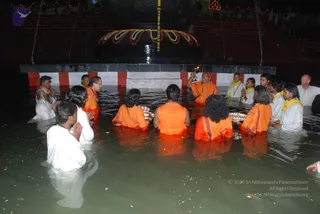A night photograph of a ceremony in shallow water in front of a large black linga-shaped monument decorated with a yellow and orange floral garland and string of marigolds, with lit lamps along the tiers behind. A man in an orange robe and gold necklaces at the upper right holds a lit lamp aloft while people around him stand waist-deep in the water with hands folded in prayer. Several women and men in orange robes stand in the water holding silver plates and offerings. Men in white robes and yellow scarves, some wearing mala beads, stand nearby, and flower petals float on the surface.