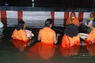 Photograph of a water ritual at dusk in a stepped tank lined with red and white painted walls. Four people in orange robes stand chest-deep in the water, seen from behind, with long dark hair and a few gold necklaces visible. On the right, a man in orange holds a silver tray of flowers and offerings. Near the top right, a man in orange holds a lit aarti lamp with a flame, and a man in a white shirt with a yellow shawl stands beside the steps. Floating flower petals dot the dark water, consistent with an abhishekam or aarti in a tank.