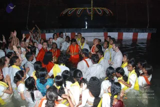 A photograph of a crowd of devotees standing waist-deep in water at night before an illuminated structure decorated with marigold-style garlands and a red-and-white striped barrier. A central figure in an orange-red robe and a long beaded mala stands in the water, holding a small brass vessel, with a garland draped around his neck; a person beside him wears a matching orange robe. Around them, many participants in white clothes with yellow and orange shawls raise their hands, some with palms pressed together, and women with long braids stand nearby. Lamps and lights glow above the water. a person is the figure in the orange-red robe with the long beaded mala at the centre of the water ritual, identified by a swept-back appearance and a distinctive mala and orange robe.