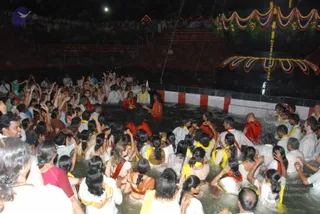 Night photograph of a large crowd gathered in a water-filled sunken tank, many people standing waist-deep in water with arms raised, some holding small objects. Several people in front wear red or orange garments and garlands, while many others wear white dhotis or sarees with yellow and orange borders. A decorated ornamental canopy with floral garlands and red-and-white painted trim stands at the upper right, and tiered stone steps with small lamps line the background under a dark night sky. The scene suggests a ritual bathing ceremony in the water tank.