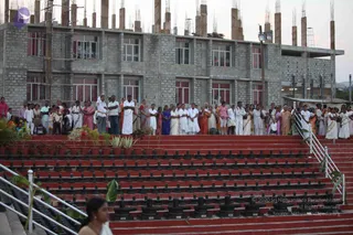 A large group of devotees stands in rows on a red-tiled stepped platform lined with black Shivalinga stones, facing away toward an unfinished grey concrete building with rebar columns and barred windows. Many wear white dhotis and kurtas or white sarees, while women wear sarees in pink, blue, orange and red. Potted plants sit along the left edge, and a woman in a white sari is partly visible in the lower foreground. The scene appears to be an outdoor ashram gathering at dusk.