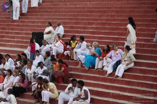 A photograph of many devotees seated on tiered red-brick steps at an outdoor ashram gathering. Most are dressed in white, with some in pink, orange, yellow and teal saris. Several women wear white and orange lanyards with name badges and colourful rosette ribbons. A woman in a white sari stands at the upper right on the steps, while a black speaker box sits on the steps at the top left. Participants sit in conversation and watch the proceedings, with children seated among the adults.