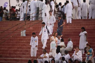 Outdoor photo of a large crowd in white clothing on red-brown stone steps. Many people in white kurtas, dhotis and salwar kameez stand along the upper railing and climb or sit on the steps. A young girl in a white outfit with a blue ribbon badge stands at left, while a man in white with a backpack and a red-and-blue rosette walks down the steps. A woman in a white dress with an orange-and-yellow garland and a blue shawl walks at centre, and a man in a white shirt with a garland sits at lower left. Several seated devotees in white wear garlands and rosettes.