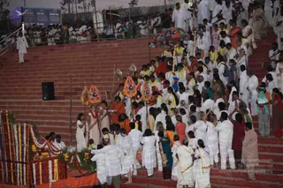 Outdoor evening procession on red brick steps at a temple-like ghat. A crowd of devotees, mostly dressed in white with some in yellow, orange and red, fills the stairs. In the centre, a man in an orange-saffron robe with a long beard walks beneath a colourful umbrella-style canopy held on tall poles, with ceremonial gold standards and decorated flags with red and blue motifs around him. To the left, a woman in white with a red shawl stands near a platform decorated with orange and yellow marigold garlands and flower strands. Many people in white kurtas and saris move up and down the steps, and a large drum is visible at the far right edge. A blue sign with white text is on the upper left wall.