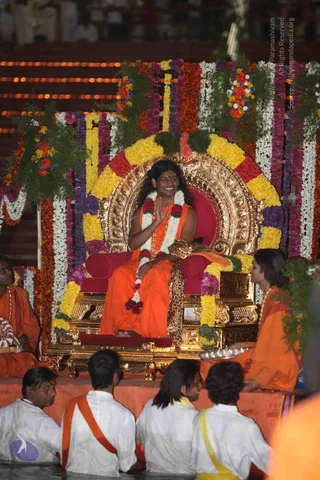 A seated figure on an ornate gold throne with a red cushion, draped in orange robes and a white and magenta flower garland, smiling and raising one hand in greeting. Around the throne are arches of marigolds, roses and greenery in yellow, red, purple and white. Flanking the throne, two people in orange robes sit on the raised platform, one holding a silver tray. In the foreground, four people in white clothes with orange and yellow sashes sit in shallow water. The scene is lit by rows of oil lamps in the background. Swamiji (probable): long dark hair, orange robes, rudraksha mala and ceremonial garland seated on a gold throne.