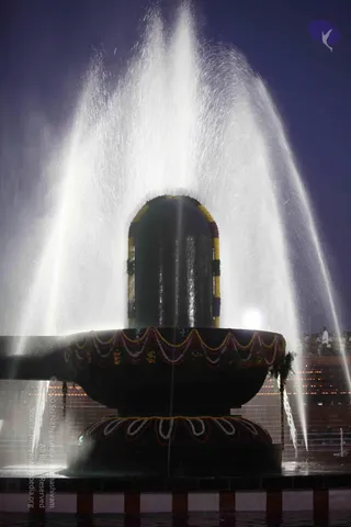 A large black linga stands on a circular yoni-shaped pedestal wrapped with garlands of white, yellow and maroon flowers, while water sprays up and cascades around it in a tall fountain against a dark blue night sky. Lamps glow faintly on a distant terrace and a few small figures are barely visible at the far right edge. The central subject is an outdoor abhishekam-style presentation of the Shiva linga under falling water.