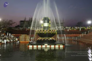 Dusk photograph of a large black Shiva linga on a tiered black peetha, decorated with a yellow-and-green floral garland and flower festoons, standing in a shallow water pool. Several fountain jets arc upward around it. Behind, rows of seated devotees in white clothes fill stone steps and a bank at the left, with an unfinished concrete building topped by many spires behind them and floodlights on either side. Strips of lamps line the pool edges. No individual person is clearly identifiable in the foreground; the crowd is seated in the background.