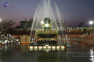 Dusk view of a large black linga monument in the middle of a water pool, with several water jets rising around it. The linga is decorated with a garland of green leaves and yellow-orange flowers at its base, and a band of striped red, white and orange is painted along the pool edge. Tiered stone steps on the right and left hold a large audience of people dressed mostly in white, seated and standing. Lights are strung along the steps and pool edge, and a partially built multi-spired structure with many small spires stands in the background under a purple evening sky. No individual person is clearly a identifiable candidate for a person in this view.