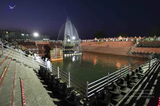 Night view of a large rectangular reflecting pool at an outdoor temple-like venue, with a tall black linga-shaped monument rising from the water's centre and a fountain jet spraying above it. Tiered stone steps on both sides are lined with rows of small lit oil lamps and seated spectators in white. Black shivalinga stones are set along the front edge of the pool. Floodlights illuminate the buildings and trees behind. A few people in white stand on a wooden platform at the water's edge near the linga, and a handful of figures are visible on the left steps. Festive lighting frames the far side of the crowd.