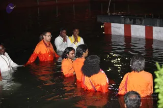 Night photograph of devotees standing waist-deep in dark water at a temple tank with steps and a red-and-white painted wall at right. Several people wear orange-saffron robes; one man in front-left wears orange with a rudraksha-style mala and glasses, and another woman in white holds his arm. Two men at the back, in white garments, press their palms together in anjali. A woman with a flower garland stands in orange. Small lights sparkle on the water surface.