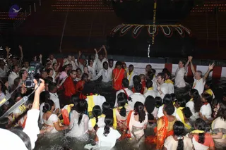 Night ceremony in a water tank with a large illuminated linga structure and garlands in the background. A central figure in a red-orange robe with a garland of flowers stands waist-deep in water, arms raised, surrounded by devotees dressed in white, many wearing yellow or orange-yellow shawls. Some participants raise their hands skyward, and a man in a red robe stands to the left. A cameraman with a camera on a tripod is visible at the lower left. Crowd of several dozen people in the water.