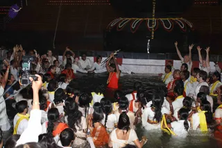 A night photograph of a crowd standing waist-deep in water at a temple tank in front of an illuminated structure. A woman in a red sari with gold necklaces stands centrally in the water holding an upright lamp or torch raised overhead. Around her, devotees in white clothes with yellow shawls and orange or red garments raise their arms and hands in devotion. A man in an orange-red garment and a priest-like figure stand nearby. A camera operator on the left films the scene with a handheld camera. Above, a decorated arch of marigold garlands and white-and-red floral ornaments hangs over the water, with a vertical garland strand suspended above.