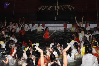A photograph of a large crowd gathered in shallow dark water at night around a central figure in a red-orange garment and a red-orange garland, who holds a guitar-like stringed instrument. Many devotees in white and yellow-scarved clothing, some in saffron and red saris, stand waist-deep, arms raised or hands folded. Behind them a dark stone structure with a tiered flower or leaf-patterned decoration and a hanging garland rises above the water, with a small fountain spray.