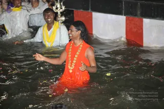 A photograph of a man standing waist-deep in dark water at night, smiling and gesturing with his open hands. He has long, dark, swept-back hair and wears a flowing saffron-orange garment with a gold-bead necklace. Behind him, a man in a white shirt with a yellow shawl also stands in the water, smiling, and several people in white clothing stand near the edge. Petals and flowers float on the water. A white and red painted wall and a silver trident are visible at the pool's edge. Appearance-based candidate, a person, is bathing in the water with devotees.