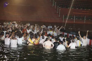 A large group of devotees stands waist-deep in a dark tiled water tank at night during a ritual bathing event. Most wear white clothes that cling to their bodies, while some women wear yellow-bordered saris and others wear red or dark tops. Many raise their arms and hands upward in devotion. A tall metal trident stands in the water near the centre, and a fountain-like jet of water rises at the right. Red-and-white striped steps and lamp lights line the tank edges in the background. A man in white at the right raises both arms high. Several people in the crowd appear to be looking toward the centre, but no figure can be clearly identified as a particular teacher with confidence.