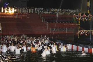 A night photograph of a crowd wading in a water tank beside a tiered red stone amphitheatre lit with rows of small lamps. Many people in white garments, some in saffron or orange cloth and yellow sashes, raise hands and palms toward a central figure holding a ceremonial staff. A tall metal pole draped with garlands of marigolds and white flowers rises from the water's edge, with a black cloth backdrop and water jets spraying behind. Spectators in white and coloured clothing sit or stand on the steps above. A balding man in plain white cloth stands in the water in the foreground. The scene reads as a mass ritual bathing or abhishekam at the water's edge.